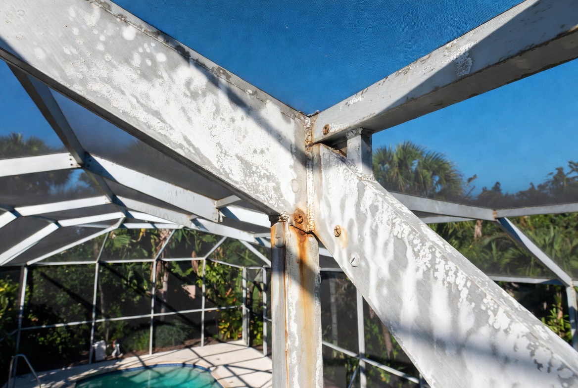 White aluminum pool cage corner with heavy oxidation, bubbling paint, and rust streaks from corroded screws above a Florida pool